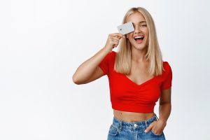 stylish-blond-girl-shows-credit-card-near-face-laughing-concept-shopping-contactless-payment-standing-white-background